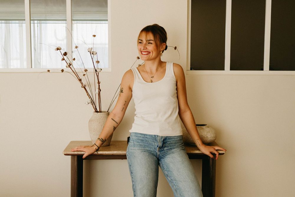 Smiling woman in a white tank top and light blue jeans leans against a wooden console table beside a vase of dried branches.