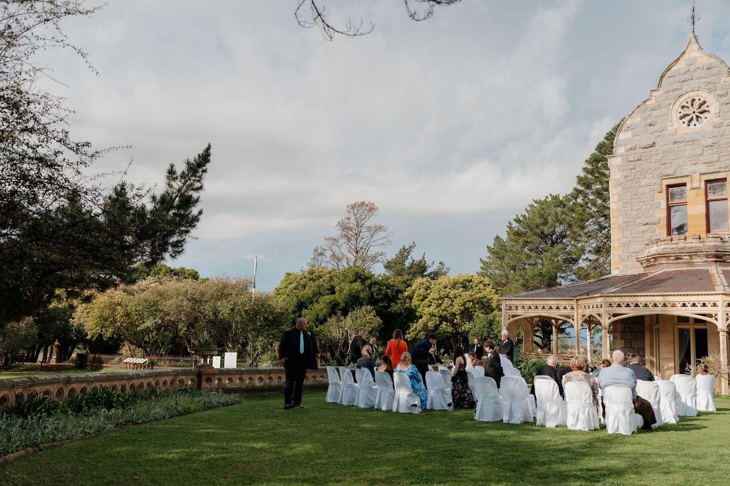 Outdoor wedding ceremony on lush green lawn beside a historic stone manor under a cloudy sky.