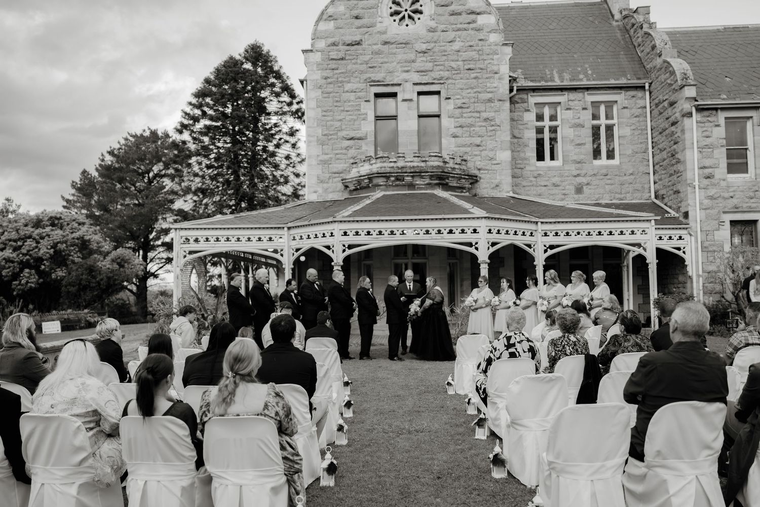 Black and white outdoor wedding ceremony at historic stone mansion with gazebo, white chairs, and bridal party.