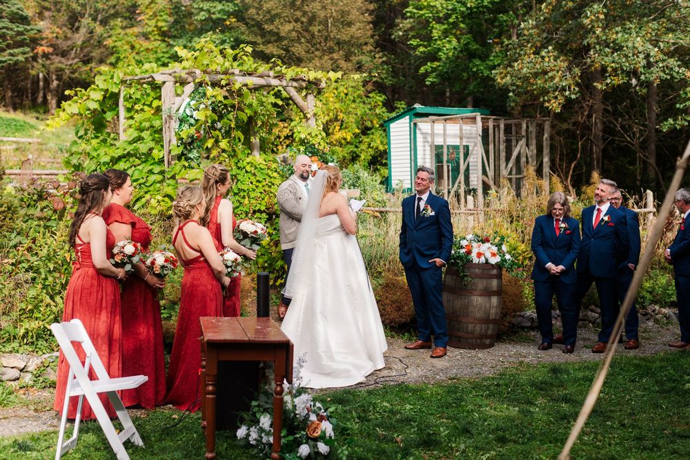 Bride and groom exchange vows at an outdoor garden wedding ceremony with bridesmaids in red dresses and groomsmen in navy suits looking on.