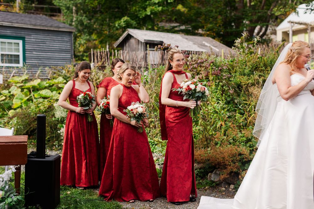 Bridesmaids in red dresses hold bouquets and smile during an outdoor wedding ceremony as the bride in a white gown and veil looks on.