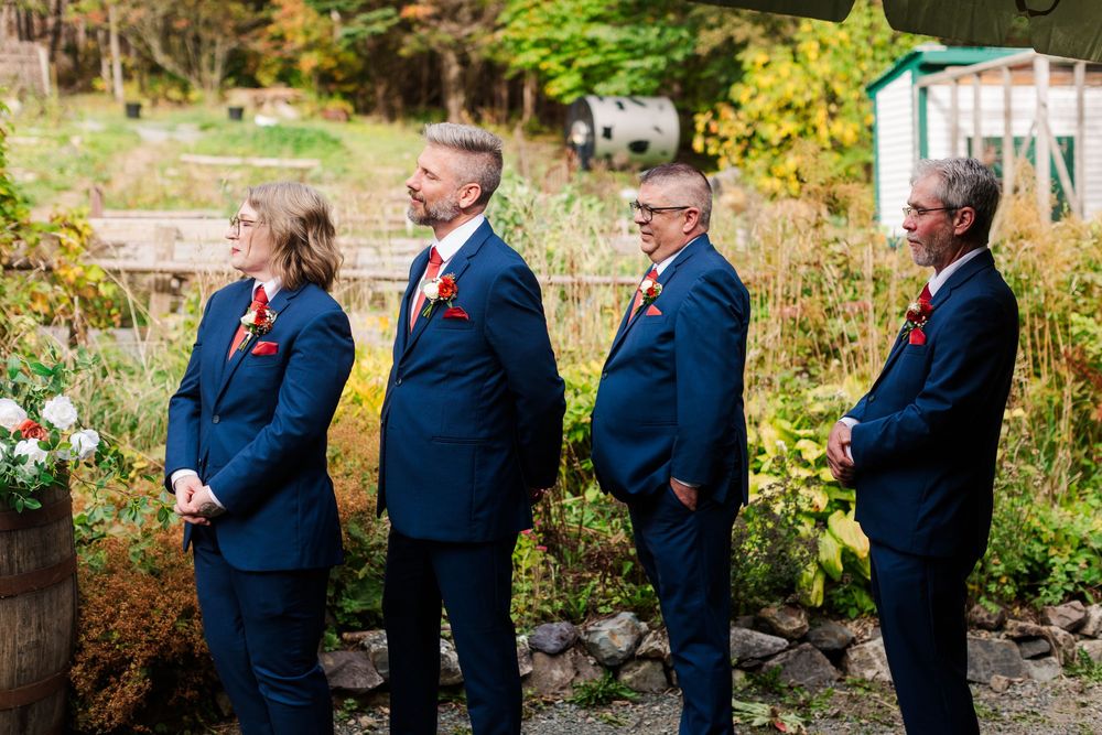 Four groomsmen in navy suits with red boutonnieres stand in a row at an outdoor garden wedding ceremony surrounded by autumn foliage.