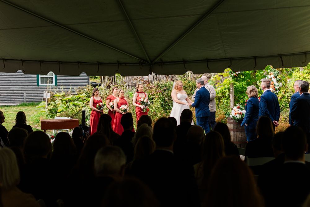 Outdoor wedding ceremony under a tent as the couple exchanges vows, with bridesmaids in red dresses and groomsmen in navy suits.