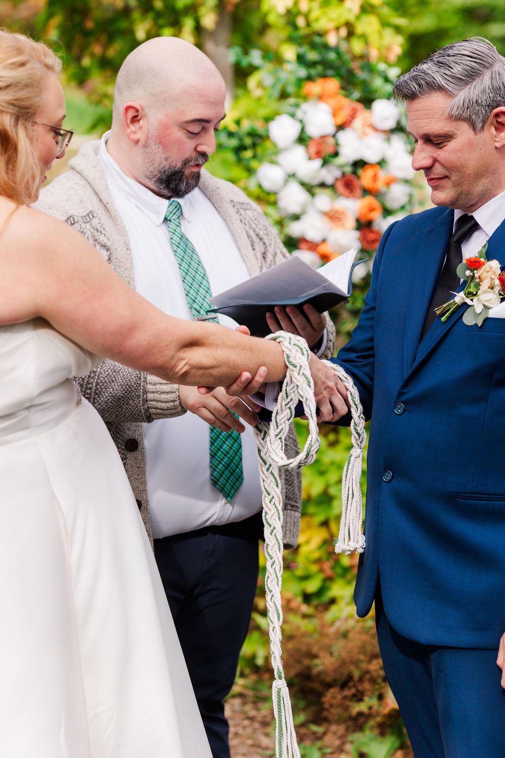 Wedding officiant in a green plaid tie performs a handfasting ceremony as a bride and groom in a blue suit have their hands bound with cord.