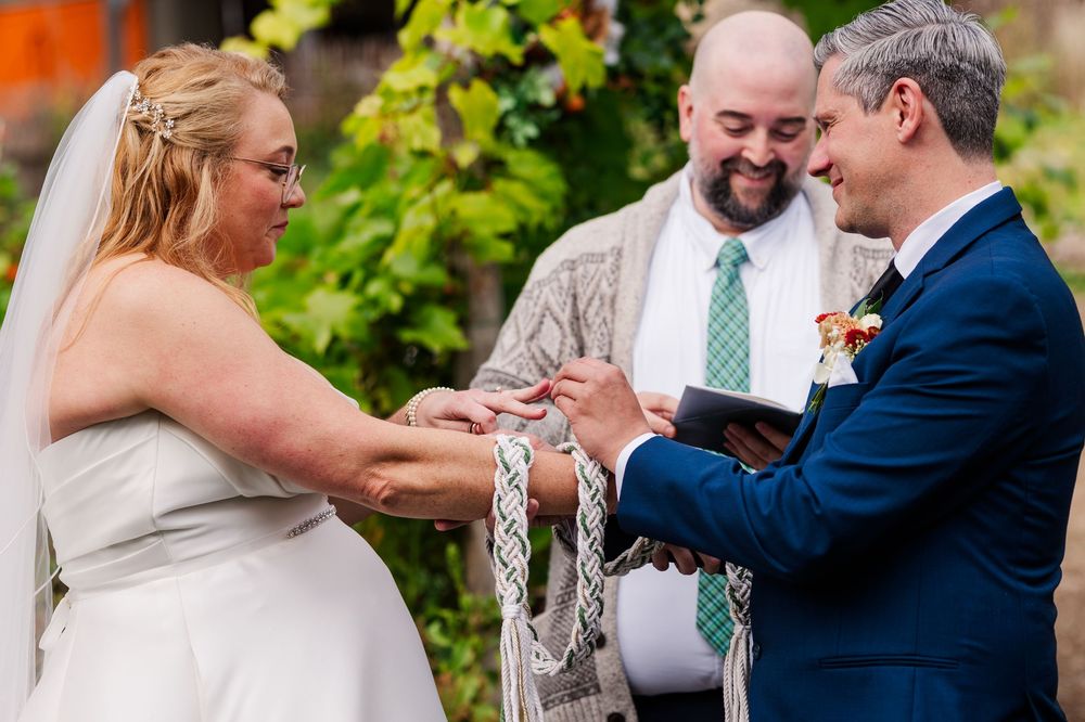 Bride and groom clasp hands bound with a braided cord during a handfasting ceremony as a smiling officiant looks on outdoors.