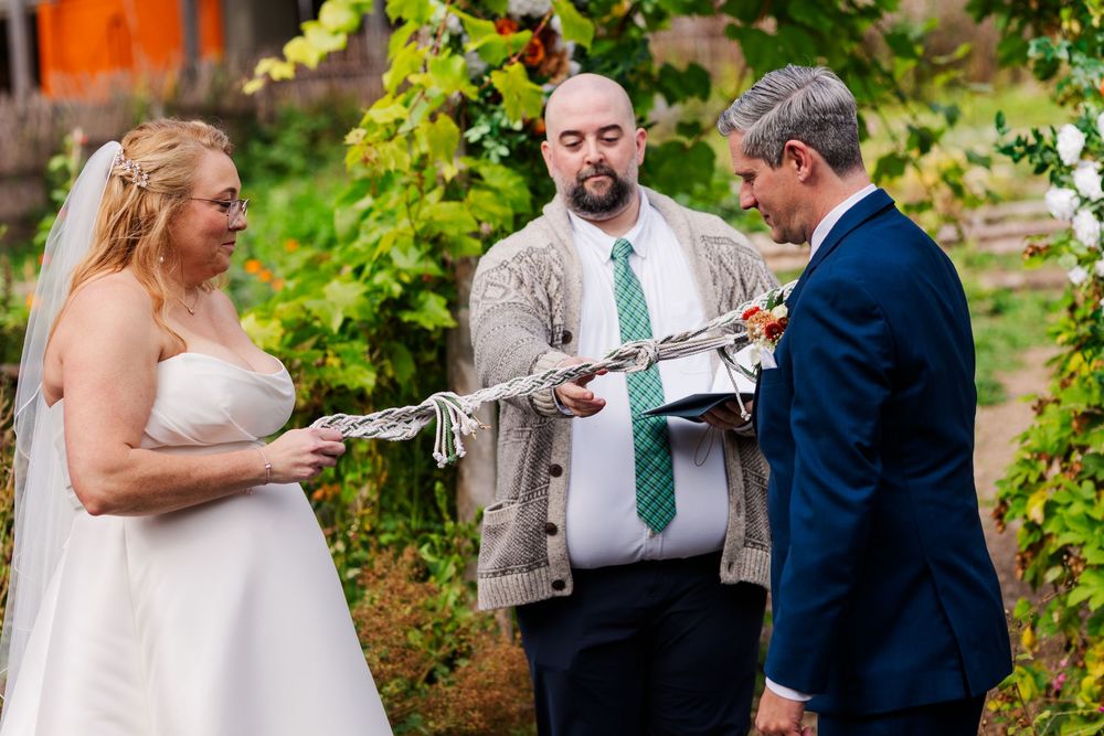 Bride in a white gown and groom in a navy suit hold a braided cord during a handfasting wedding ceremony led by an officiant outdoors.