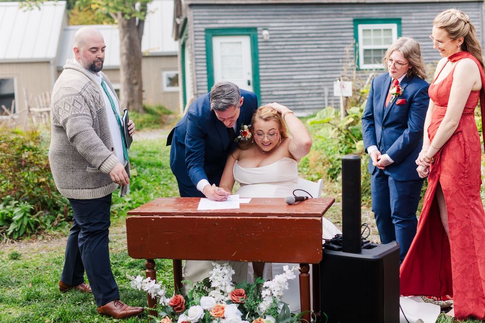 Bride and groom sign the marriage license at a wooden table during an outdoor wedding ceremony as guests look on beside a rustic building.