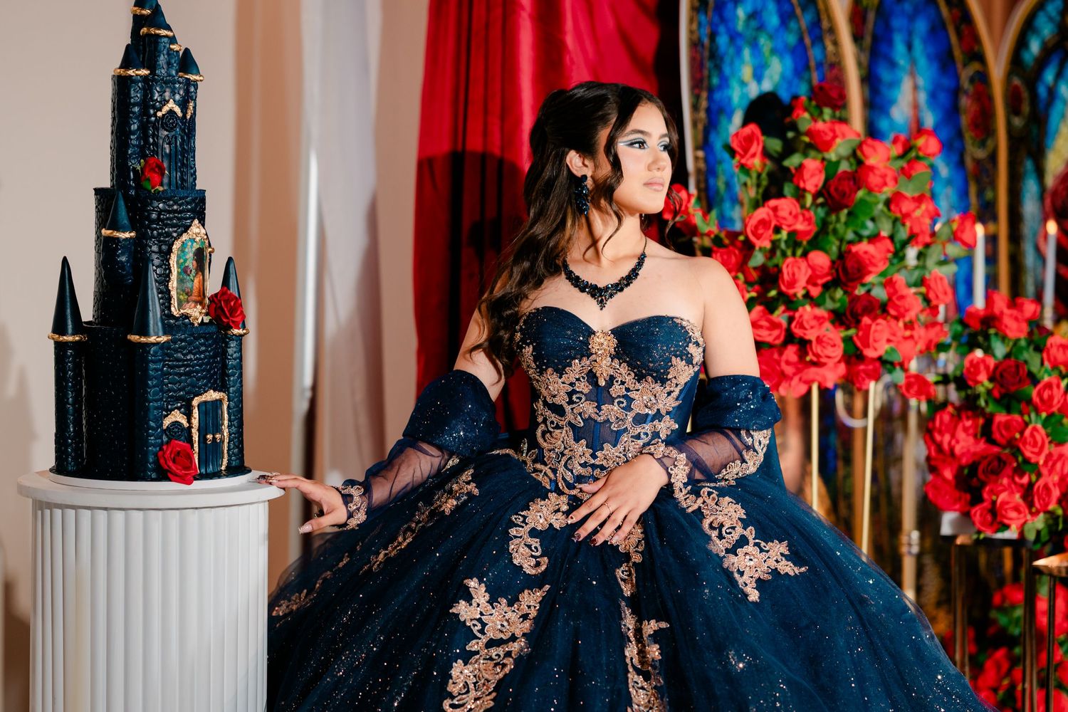 Young woman in a navy blue quinceañera ball gown with gold embroidery posing beside a castle-shaped cake near red roses and drapes.