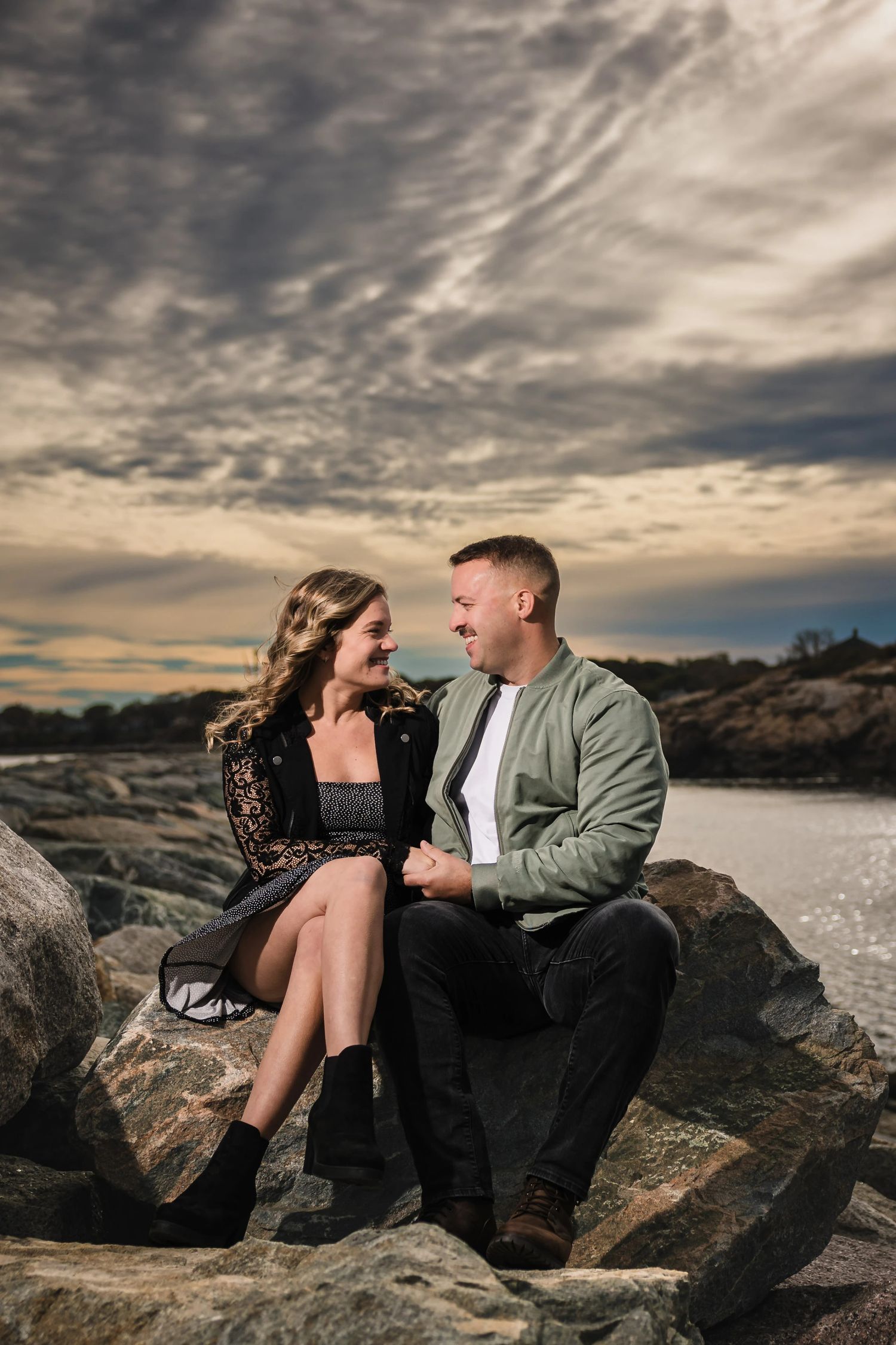 Couple sitting together on rocks by water, gazing at each at dawn. Engagement at Rockport Harbor, Rockport, MA