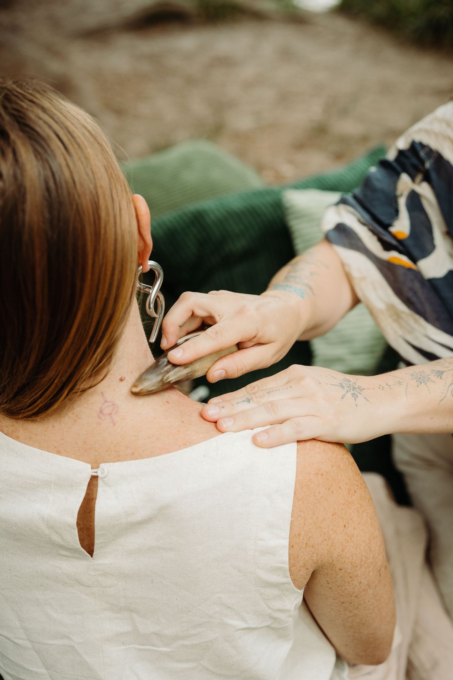 Tattooed hands performing gua sha stone massage on a woman's shoulder, wearing a white dress outdoors.