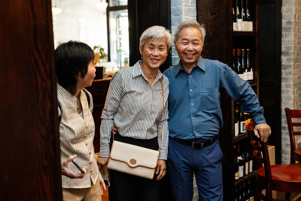 Three elderly Asian adults smiling together in a restaurant, with wine bottles displayed on shelves in the background.