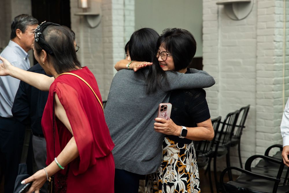 Two women share a warm hug at an indoor gathering, with guests in colorful attire socializing in the background.