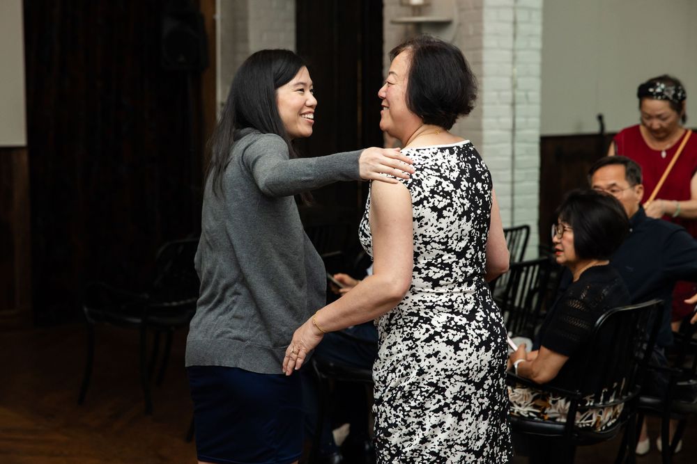 Two women smiling and embracing warmly at an indoor social gathering event.