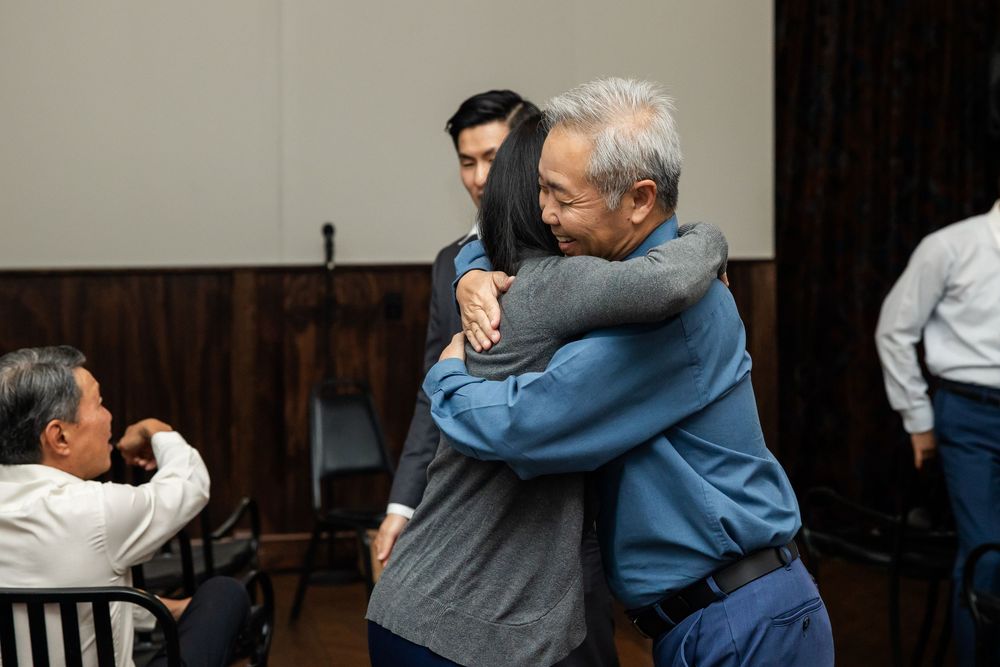 Two people share a warm hug in a room while others sit nearby in chairs.