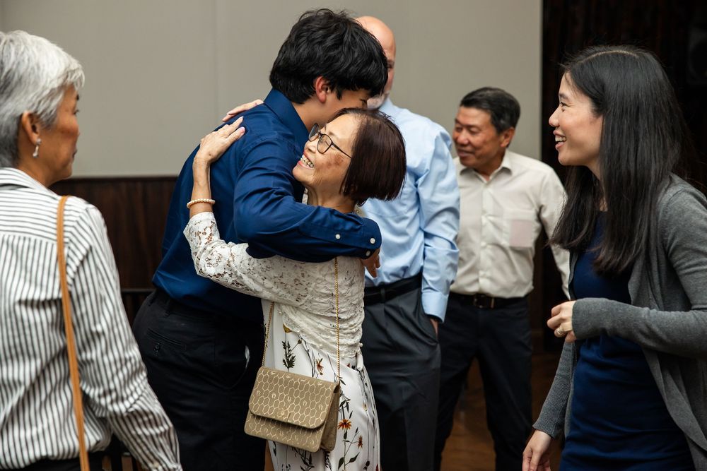 People hugging warmly at an Asian family gathering, with smiling guests socializing in the background.