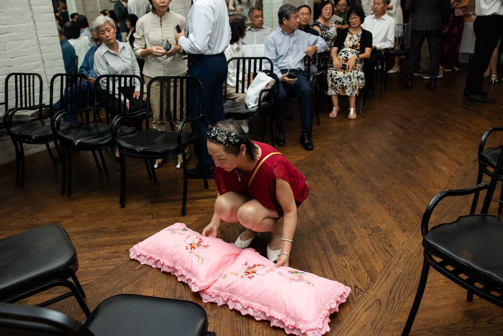 Woman in red dress arranging pink decorative pillows on wooden floor at a crowded indoor cultural event.