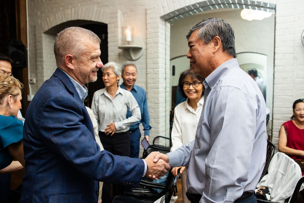 Two men shaking hands at a formal indoor gathering with guests socializing in the background.