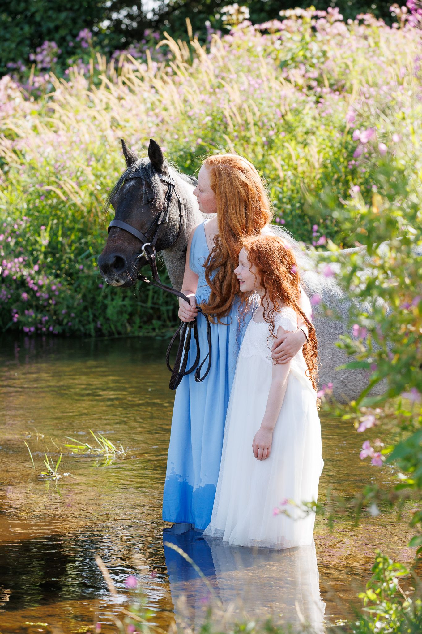 Woman walking two chestnut horses down a sunlit gravel path lined with green trees at a horse farm.