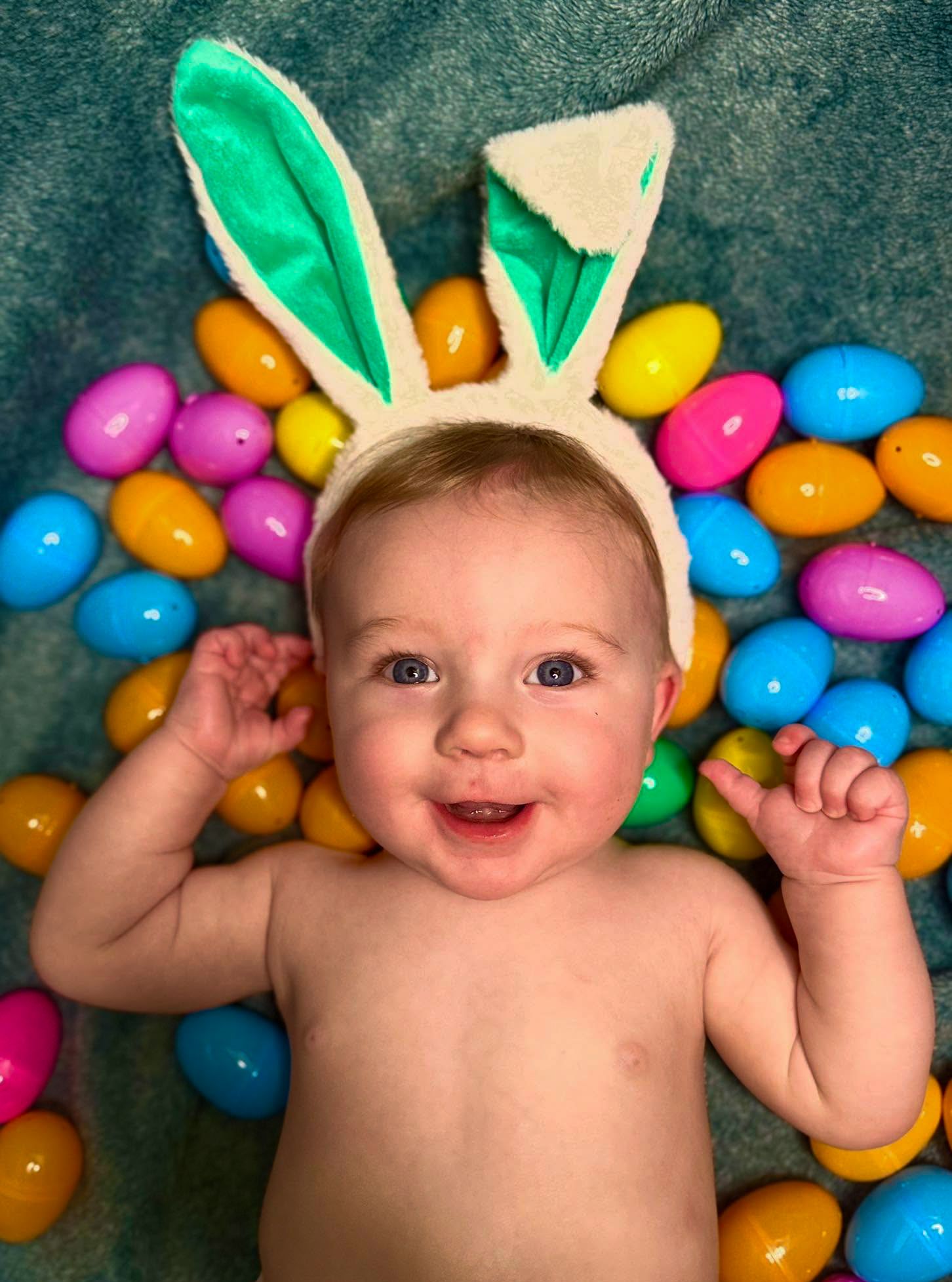 Smiling baby wearing plush bunny ears lies on a blue blanket surrounded by colorful plastic Easter eggs.