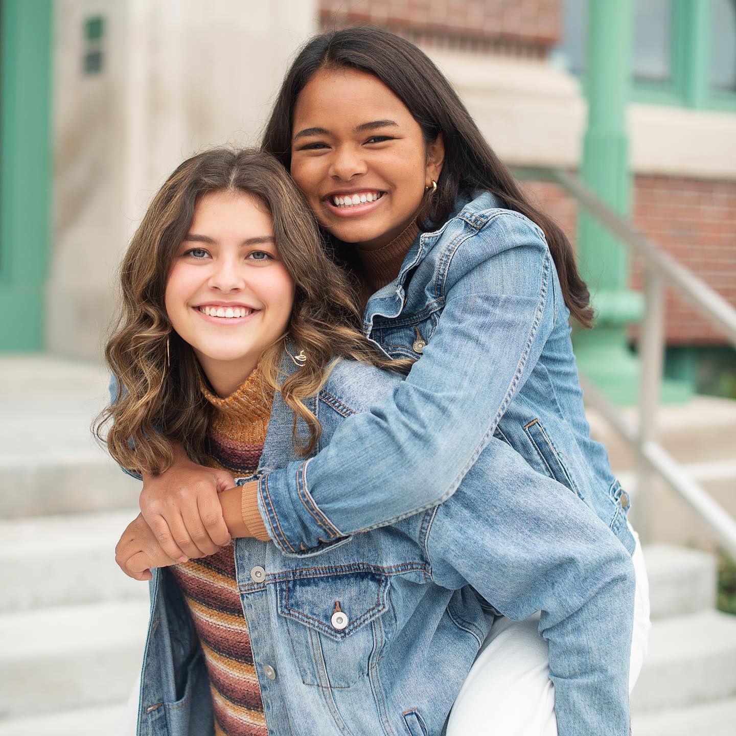 Two smiling teenage girls pose for a fun piggyback photo outside a brick building.