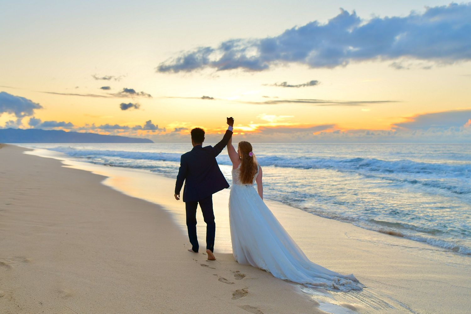 Newlywed couple holding hands and raising glasses at sunset on a tropical beach in wedding attire.
