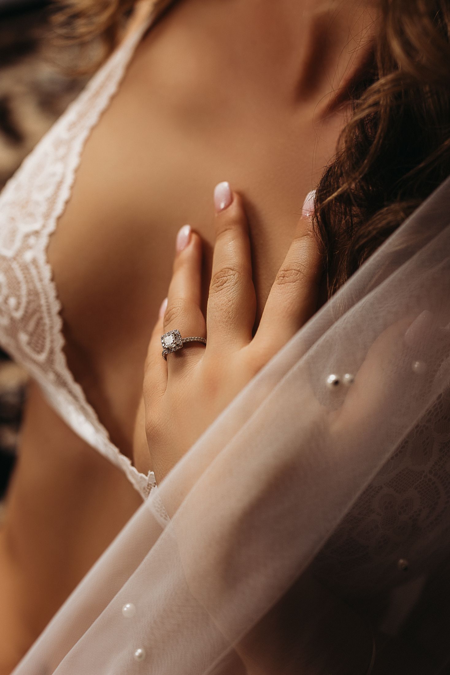 Close-up of a bride's hand adorned with a diamond engagement ring, resting on a white lace bridal gown and veil.