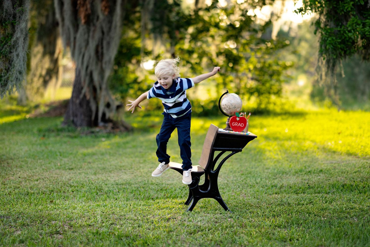 Excited blonde boy jumping outdoors near a school desk with globe and apple, back-to-school concept in a park.