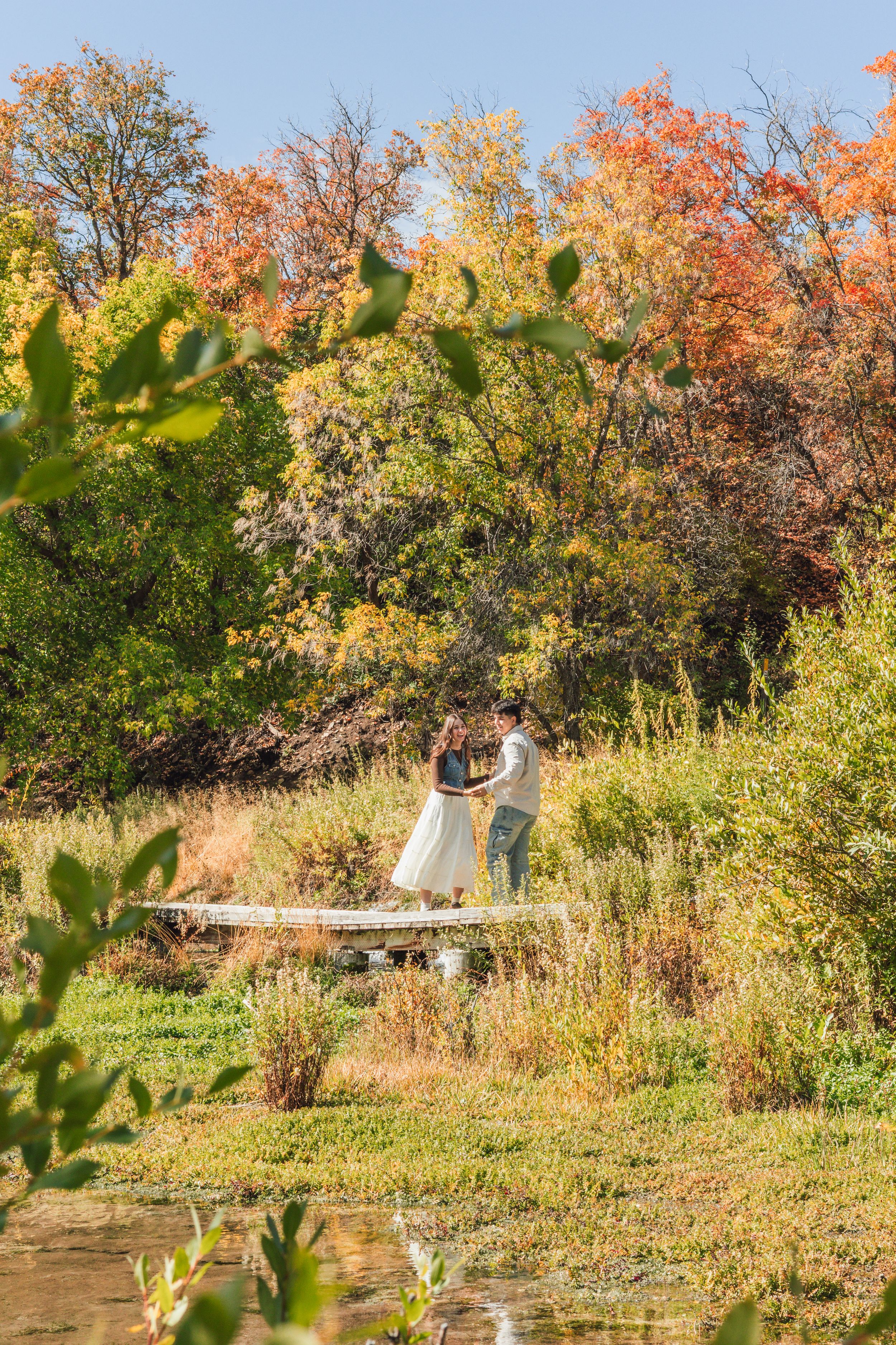 Couple embracing on a wooden dock surrounded by vibrant autumn foliage in orange, yellow, and green hues.