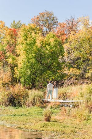 Couple standing on a wooden dock surrounded by vibrant autumn foliage in shades of green, yellow, and orange.