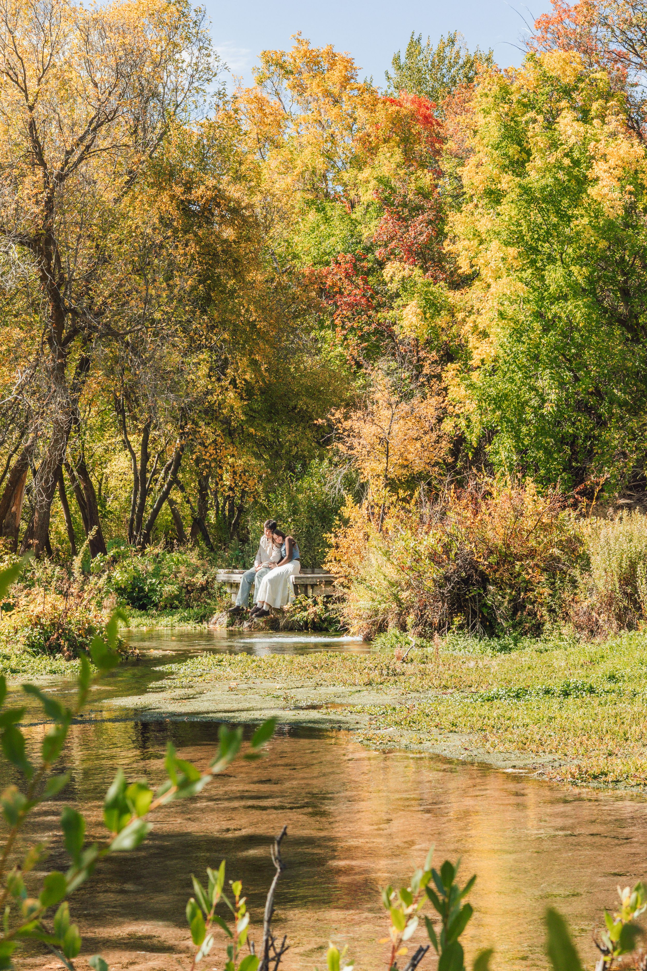 Couple sitting on a dock surrounded by colorful autumn trees reflected in a serene pond.
