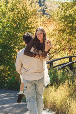 A joyful woman shows off her engagement ring while being carried by her partner on a sunny autumn trail.