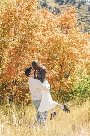 Couple embracing and kissing in a golden autumn field surrounded by vibrant orange and yellow fall foliage.