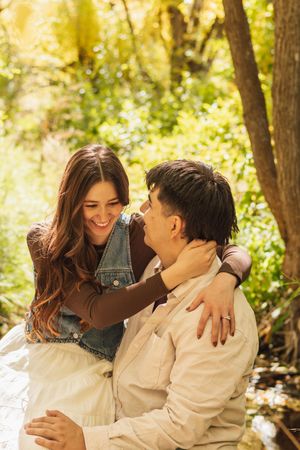 A happy couple embraces and smiles at each other in a sunlit forest setting, woman wearing denim vest.