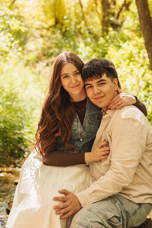 A young couple embraces outdoors in a sunlit forest, the woman in a white skirt and denim vest resting her head on his shoulder.
