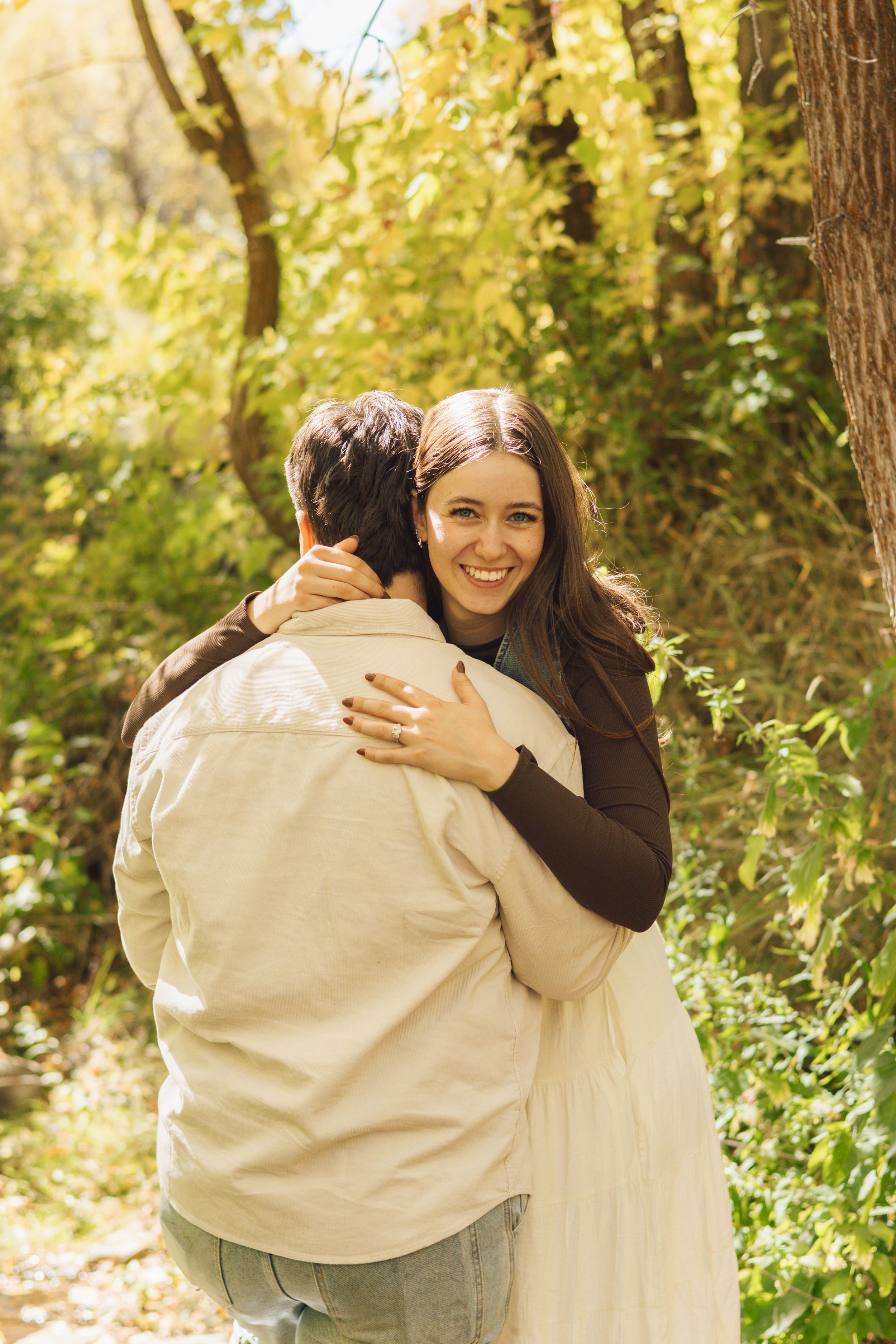 A smiling woman with long hair hugs a man from behind in a sunlit autumn forest setting.