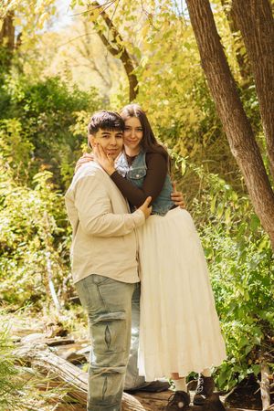 Young couple embracing in a sunlit forest, smiling warmly among green trees and autumn foliage.