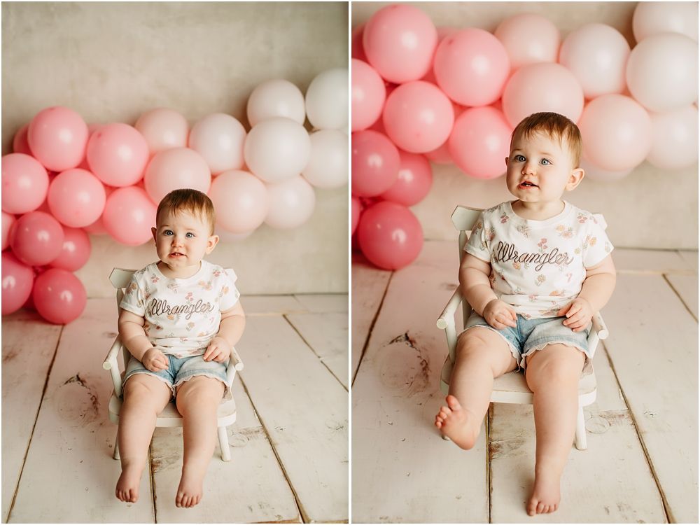 Baby girl in denim shorts sits before pink balloon backdrop during first birthday cake smash photo session.