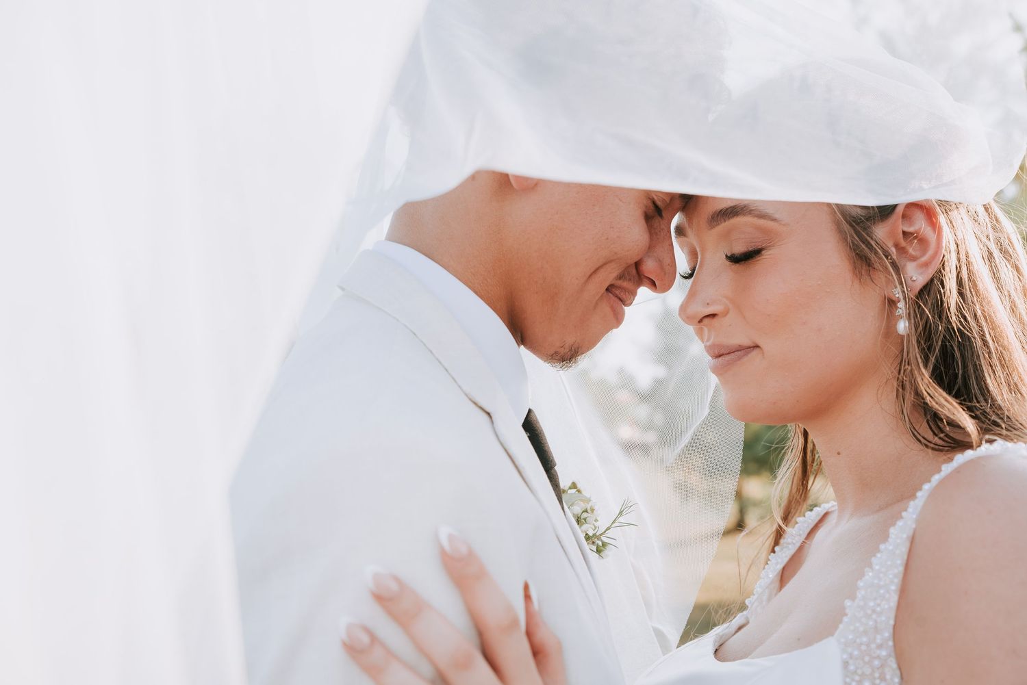 A couple in wedding attire share an intimate moment outdoors, bride holding greenery bouquet in white gown with veil.