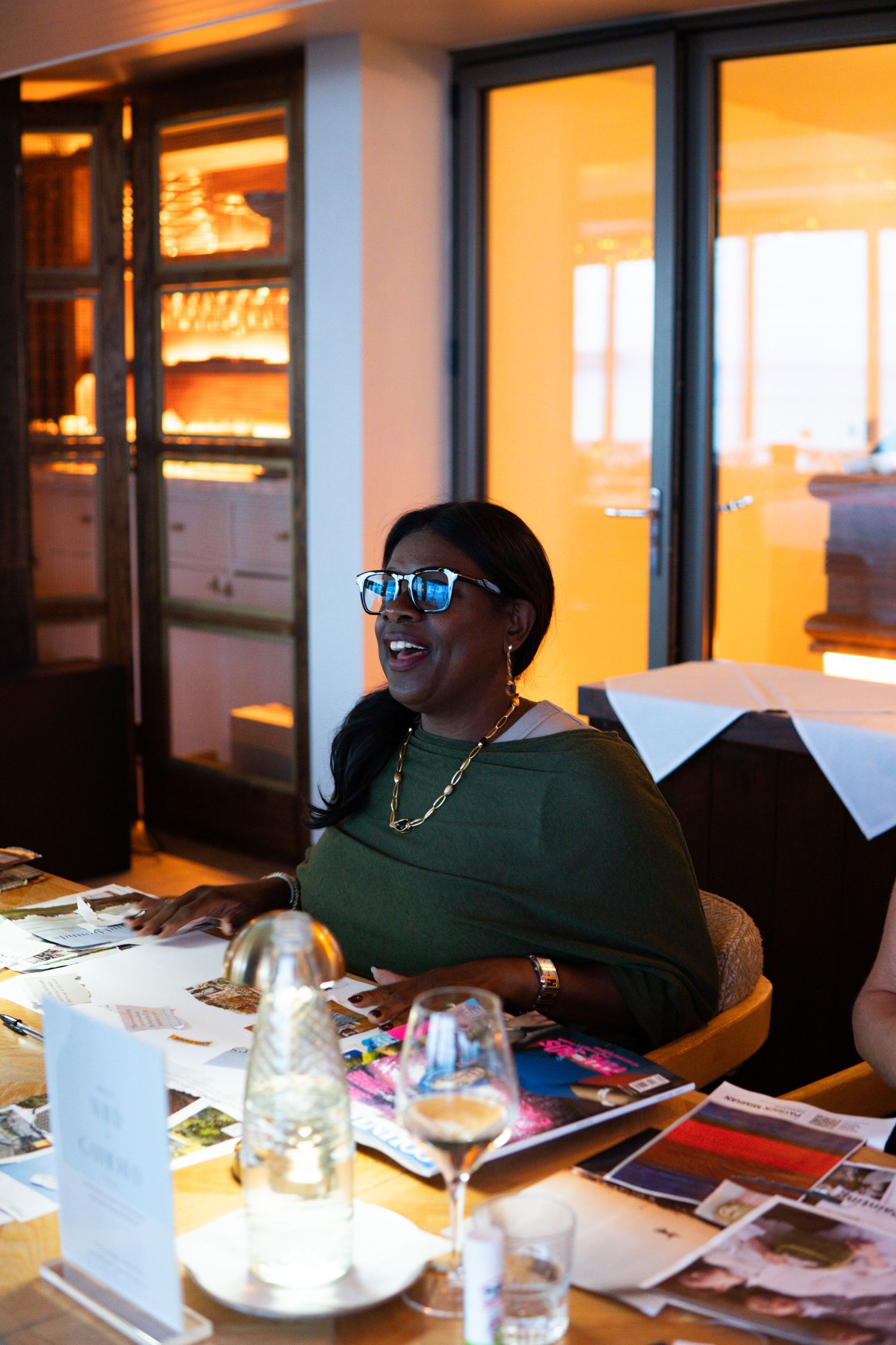 Woman in sunglasses and green top smiling at a table covered with magazines and wine glasses indoors.