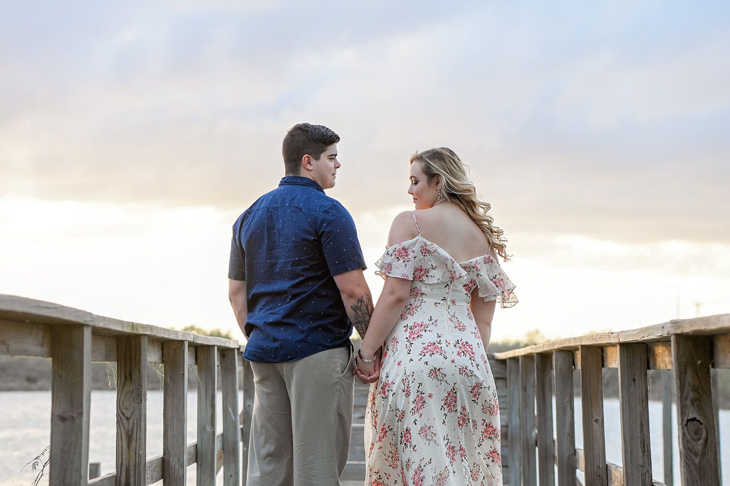 Couple holding hands on a wooden bridge at sunset, man in blue shirt and woman in floral dress.