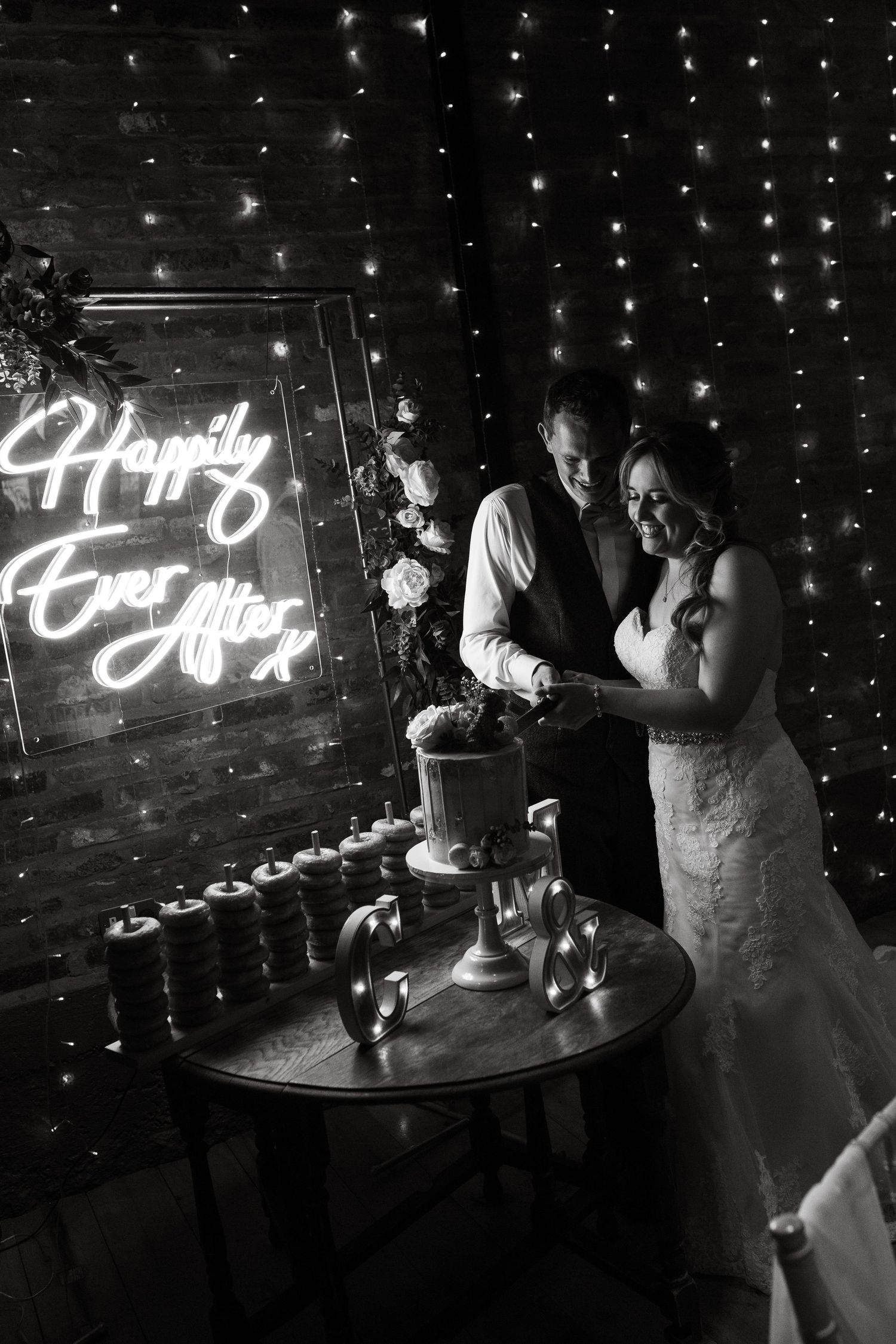 Black and white photo of bride and groom cutting wedding cake beside a neon Happily Ever After sign.
