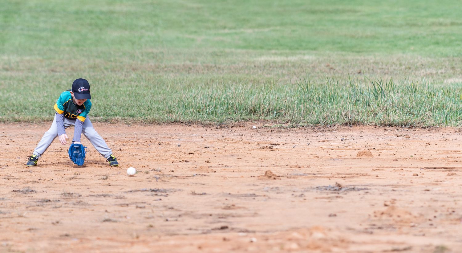Young boy in a blue and red soccer uniform and black cleats kicks a ball while running across a grassy youth soccer field.