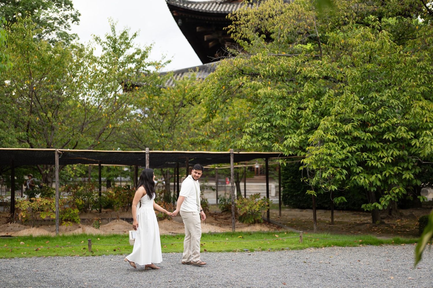 Couple in white outfits holding hands and walking along a gravel path beside lush green trees and a traditional Japanese temple building.