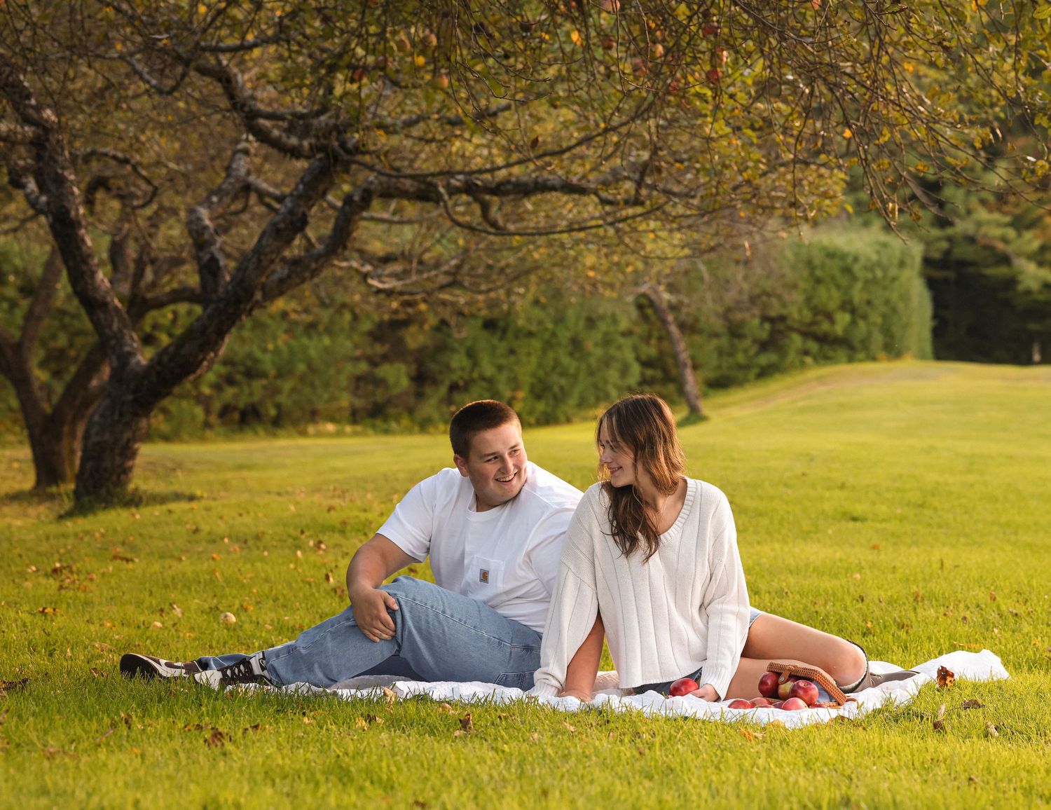 Couple sitting on white blanket in lush green park under trees, sharing a romantic moment on sunny day.