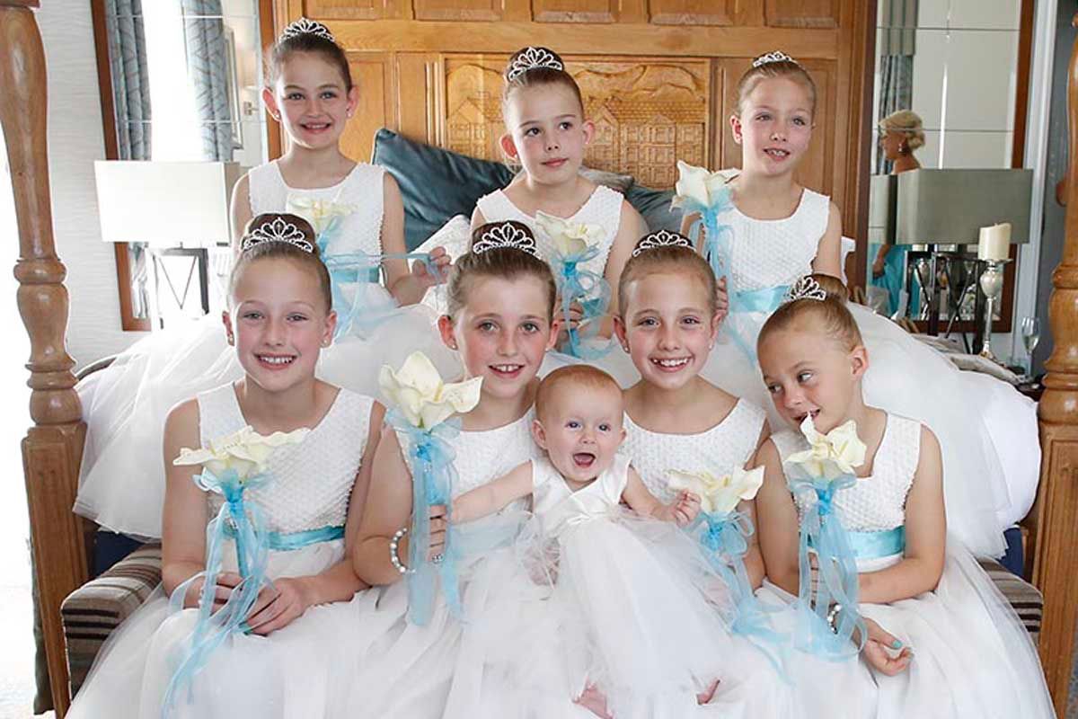 Young bridesmaids and flower girls gathered on a bed in white dresses with pale blue sashes at the Inn on the Lake