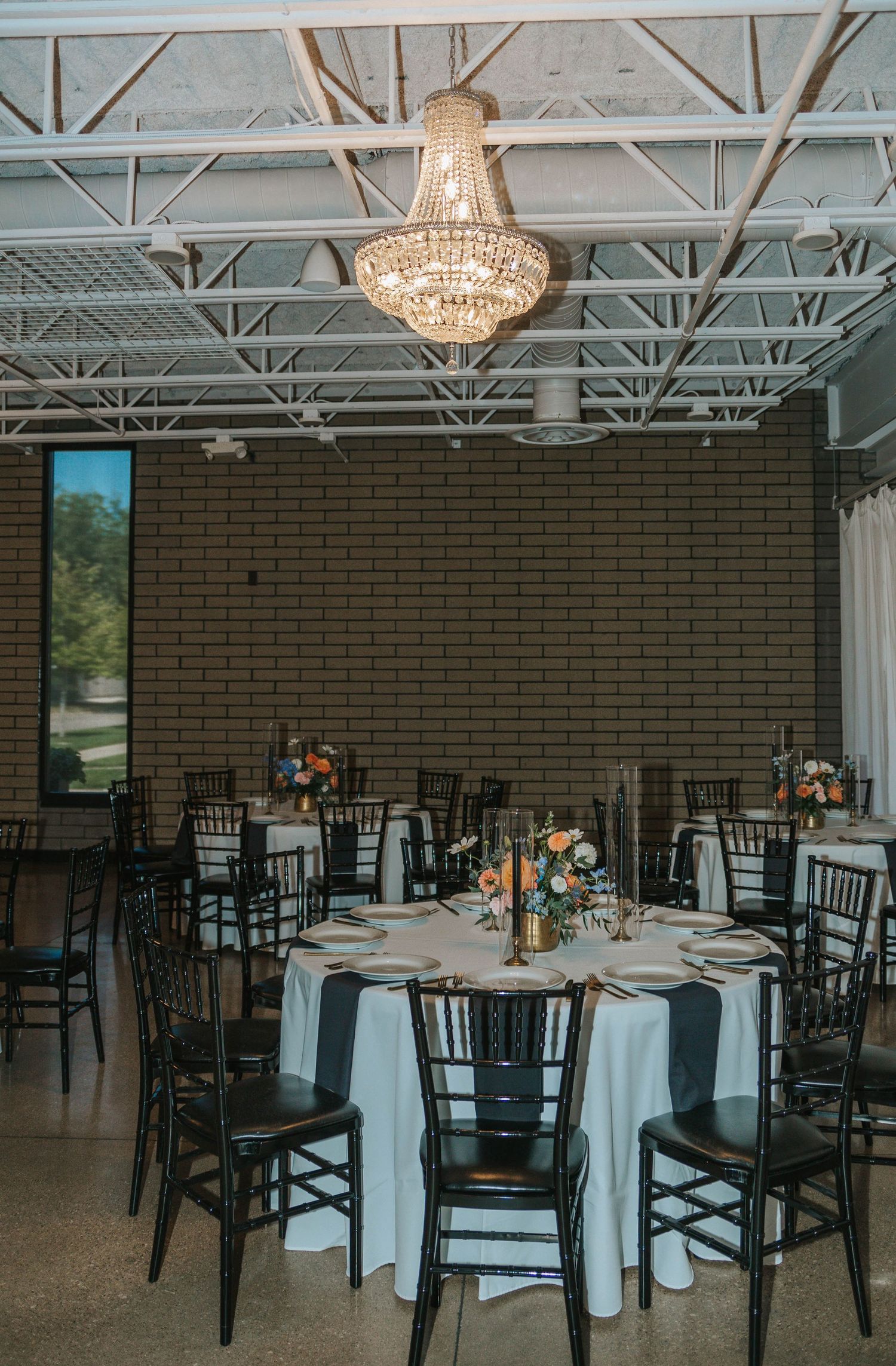 Elegant wedding reception hall with black Chiavari chairs, white linens, and colorful floral centerpieces featuring orange and blue blooms.