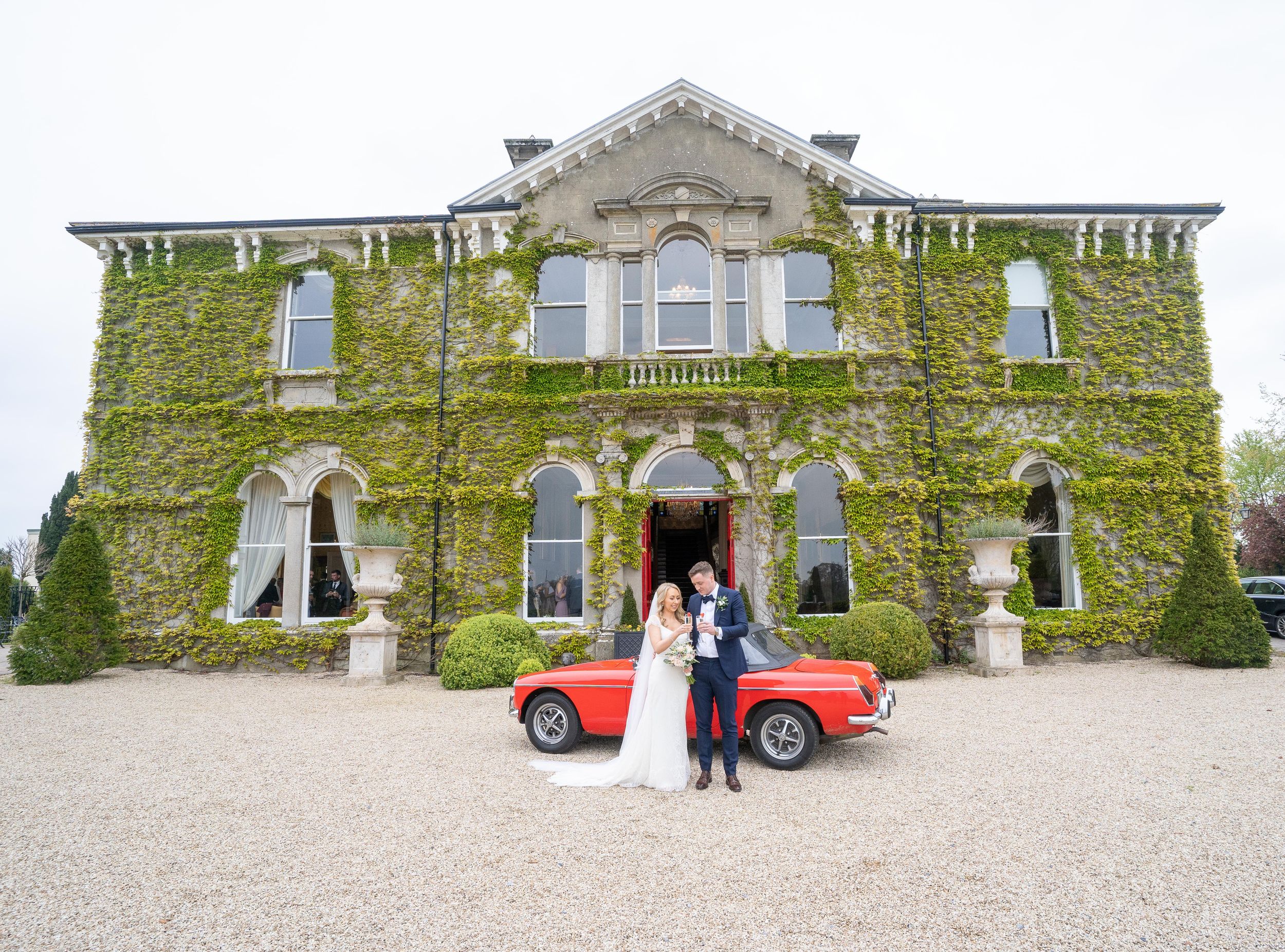 Bride and groom pose before ivy-covered Lyrath house, co Kilkenny  with classic red vintage car on gravel driveway by AK wedding photography