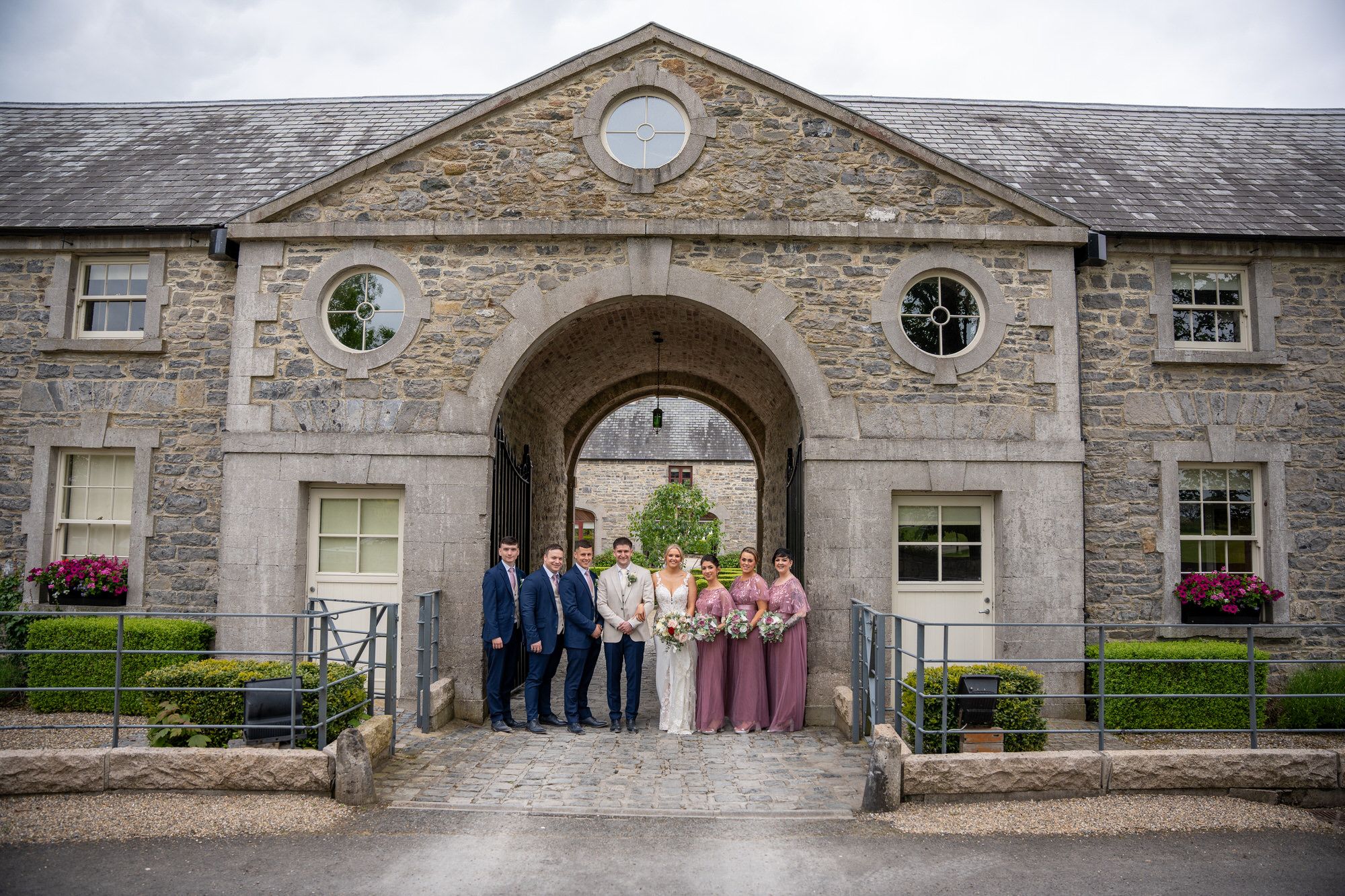 Wedding party posing under stone archway at Clonabreany house, bride in white, bridesmaids in mauve gowns Photo by AK wedding photography