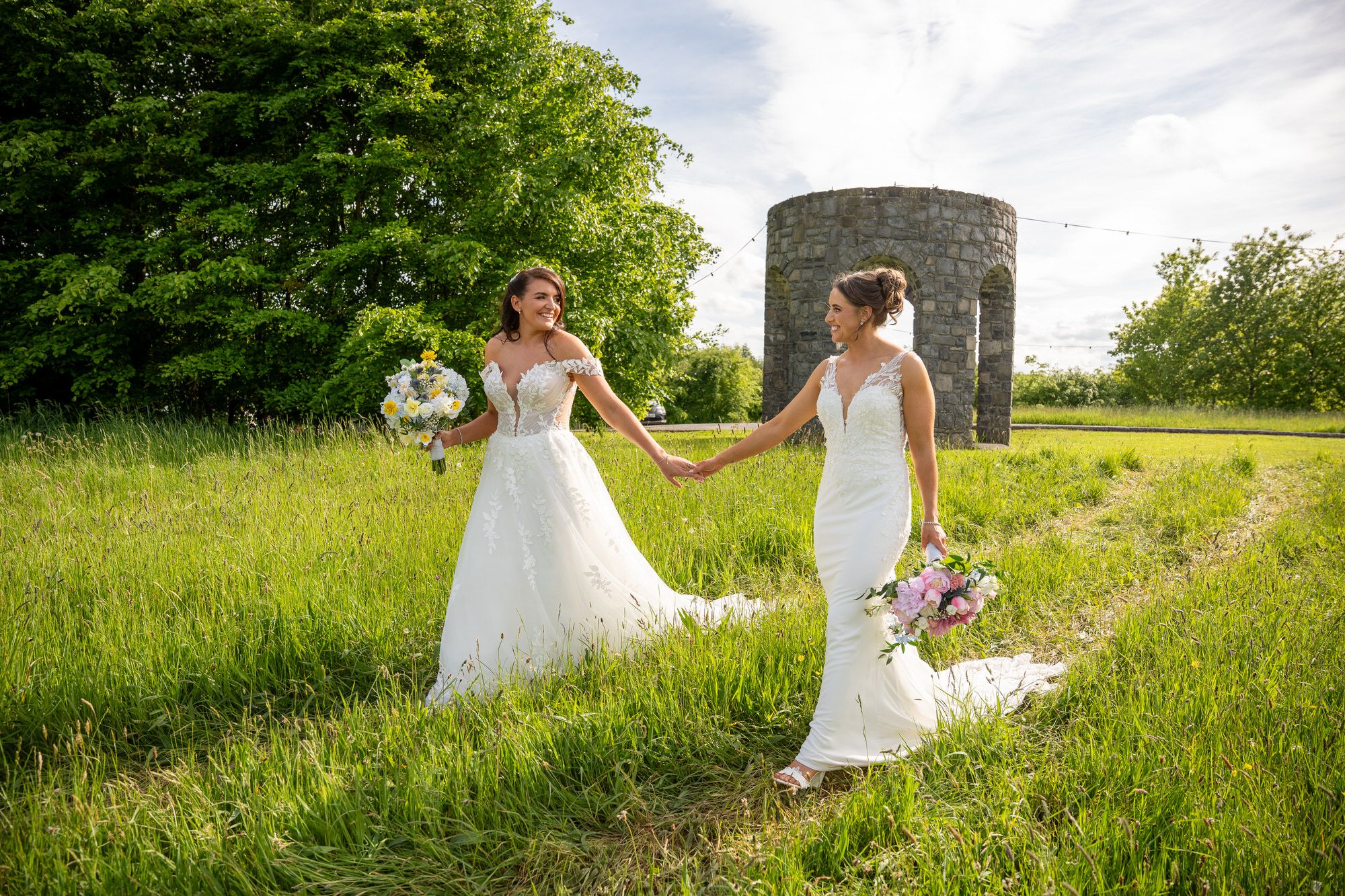 Two brides in white gowns holding bouquets walk hand-in-hand through a lush green field at Mount Druid, Westmeath photo by AK Photography