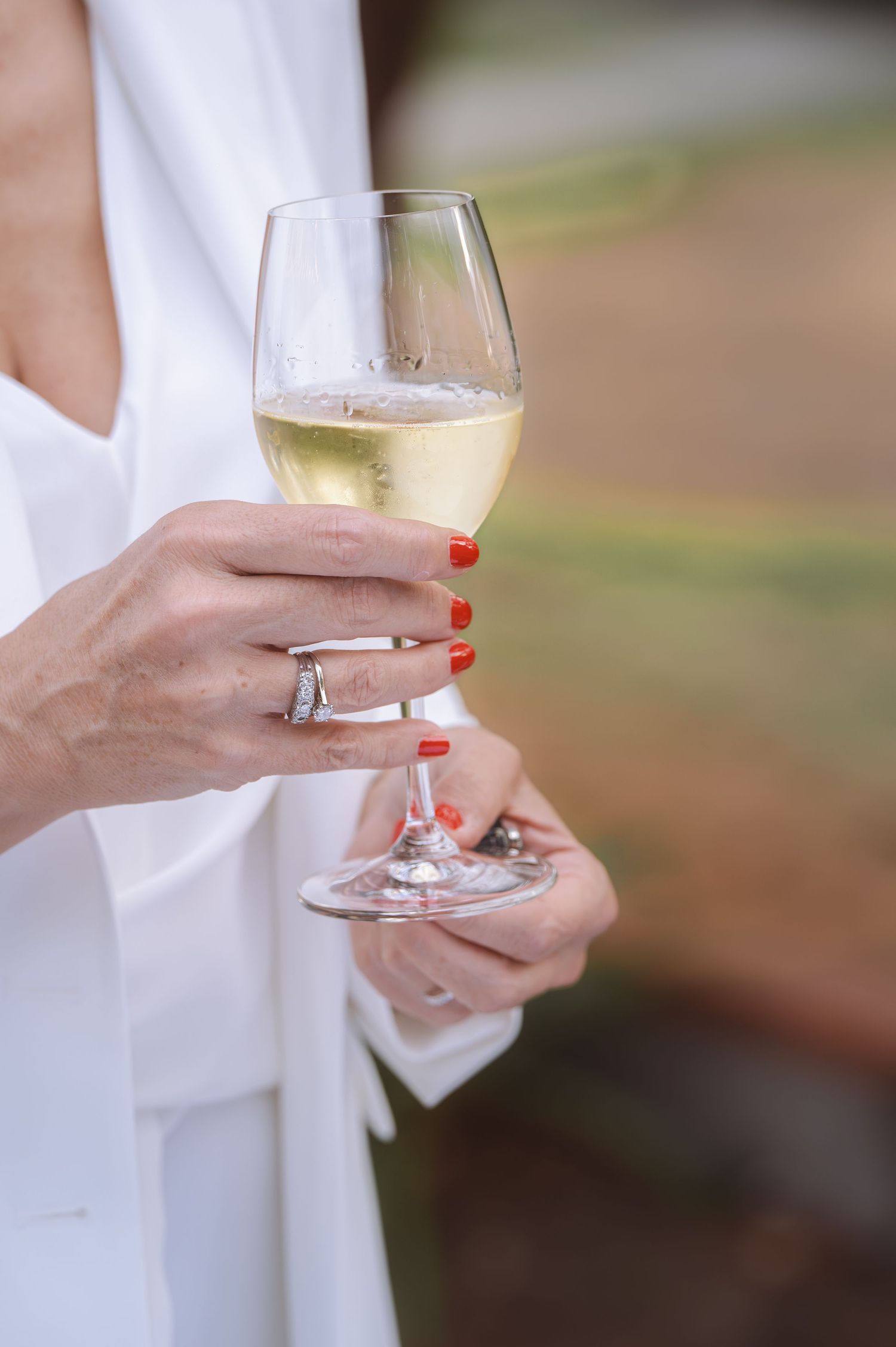 Marié en tenue blanche tenant un verre de champagne avec des ongles rouges manucurés et une bague en diamant.